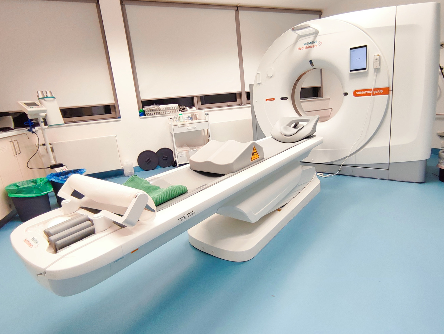 Ein modernes CT-Scanner-Gerät von Siemens in einem medizinischen Raum mit blauem Boden und medizinischem Equipment im Hintergrund.